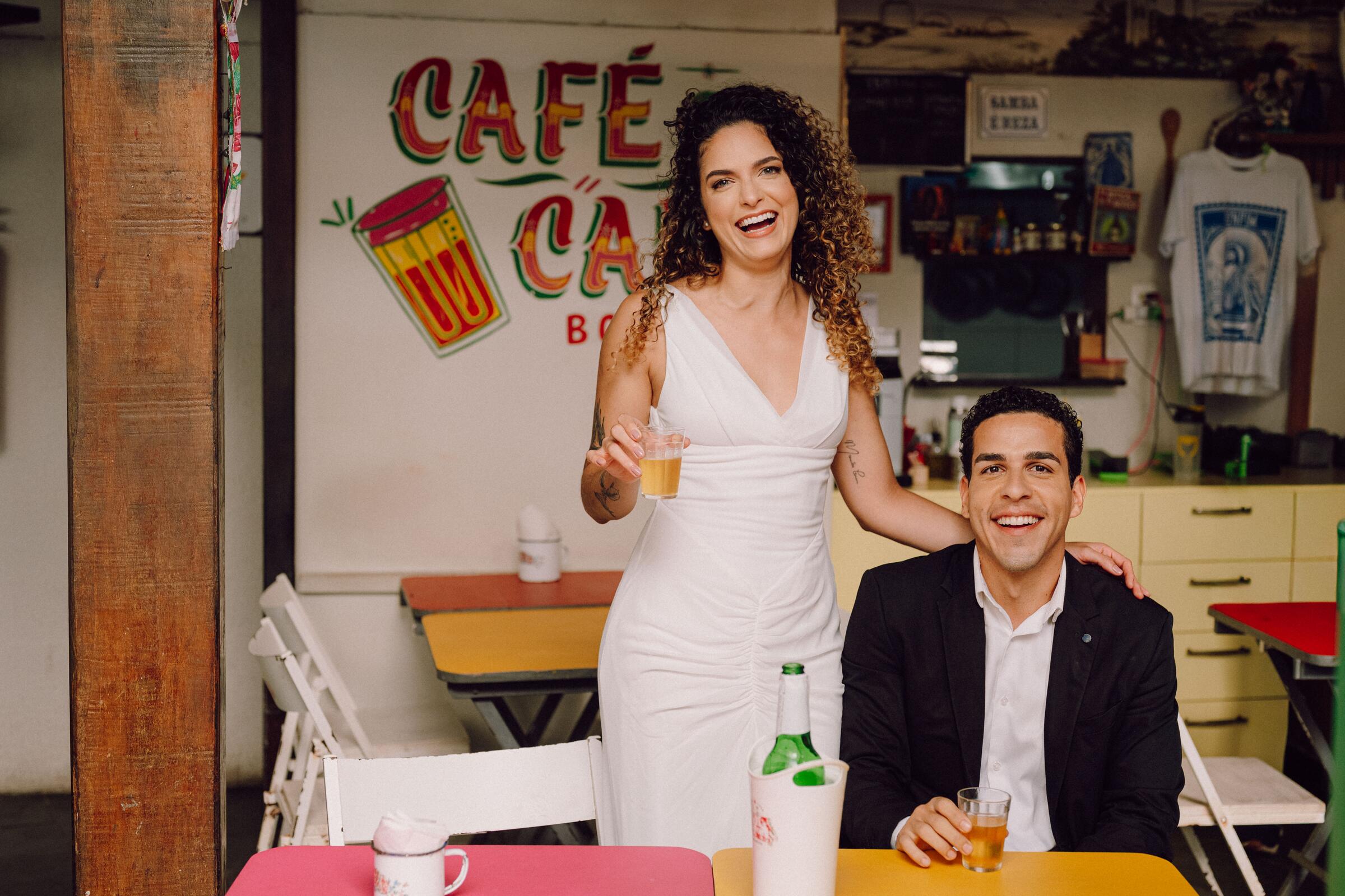 Tai brindando em pé ao lado de Leo, sob a placa Café e Cana Boteco