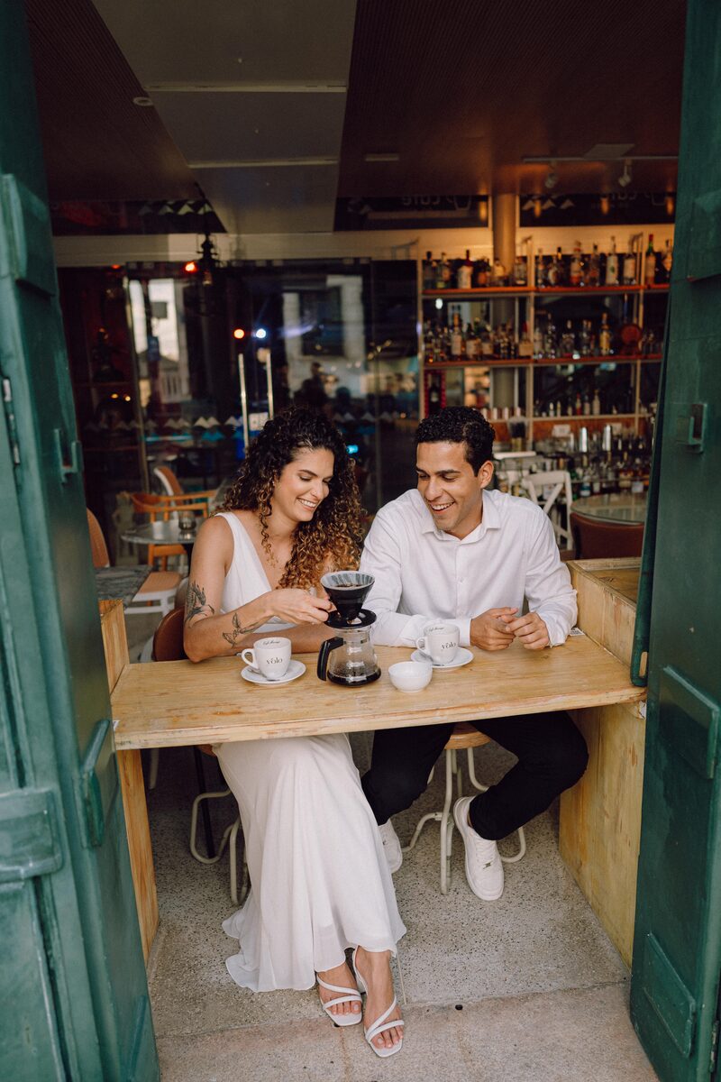 Leo e Tai tomando café em mesa de boteco, copo americano na mesa