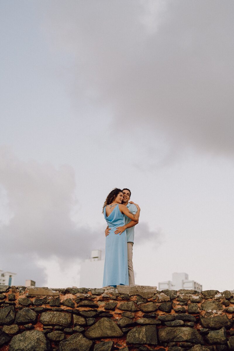 Tai de vestido azul, braços abertos, em cima de um muro de pedra