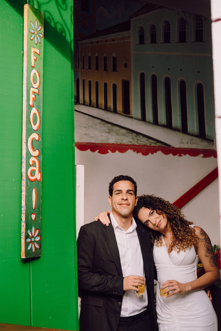 Leo e Tai abraçados e sorridentes em frente à fachada colorida do Fofoca, copo na mão