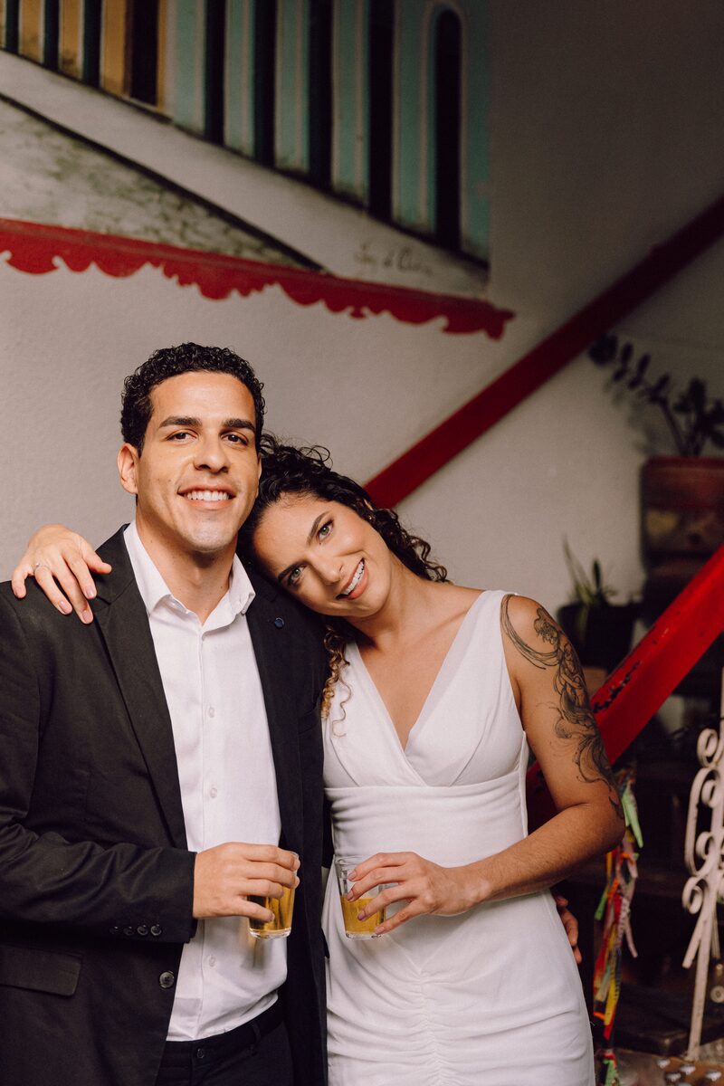 Leo e Tai sorrindo, taças na mão, em uma escadaria colorida